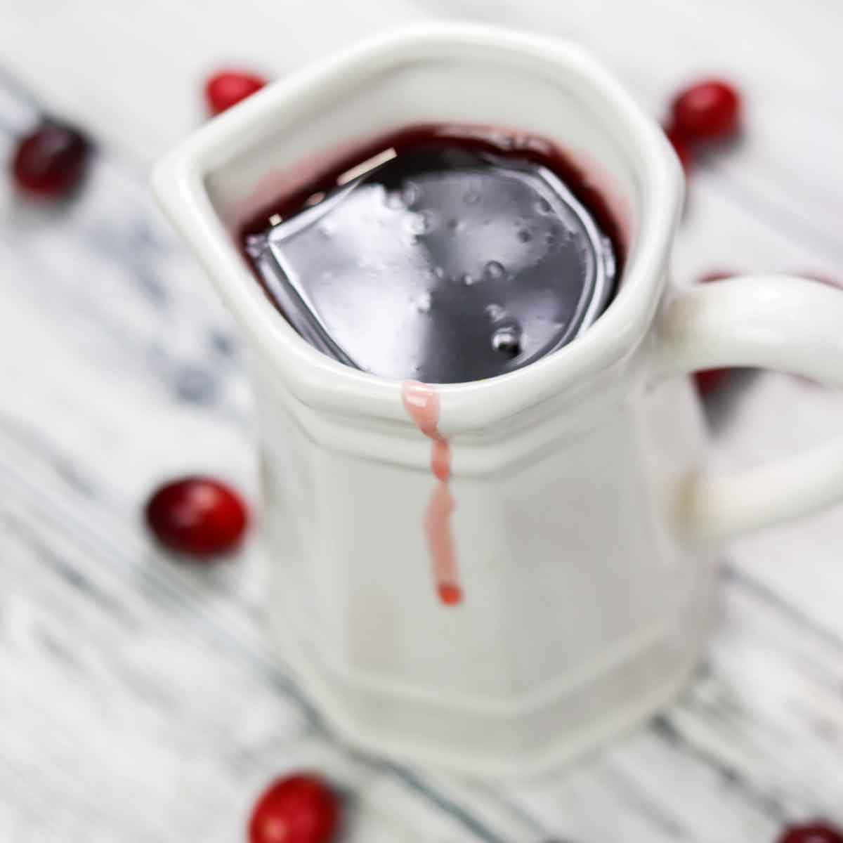 cranberry syrup in a white pitcher with a white wood background and cranberries strewn about